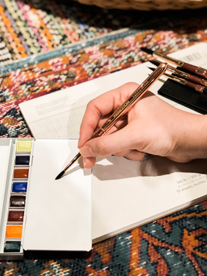A hand holding a paintbrush on a half pan palette, paper pad and paintbrushes on a carpet.