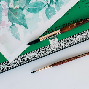 Two paintbrushes on a white background with a pad of paper and a painting