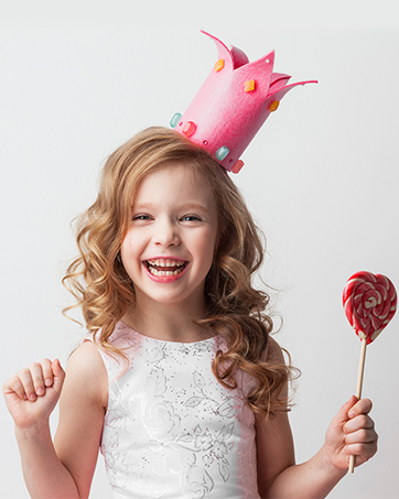 Smiling child wearing a white dress and pink crown-shaped headpiece, holding a red lollipop against a white background.