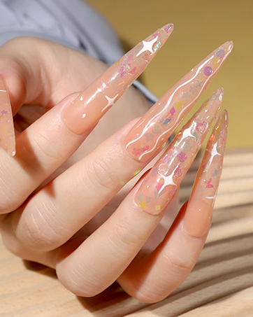 Close-up of long, pointed acrylic nails with transparent and white swirl designs. Nails are shown on a hand resting on a wooden surface.